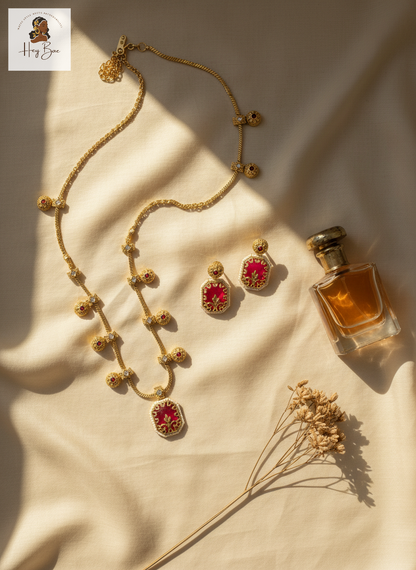 Hey Bae ruby red and gold geometric necklace set with red stones and earrings on a beige fabric background with a bottle of perfume.