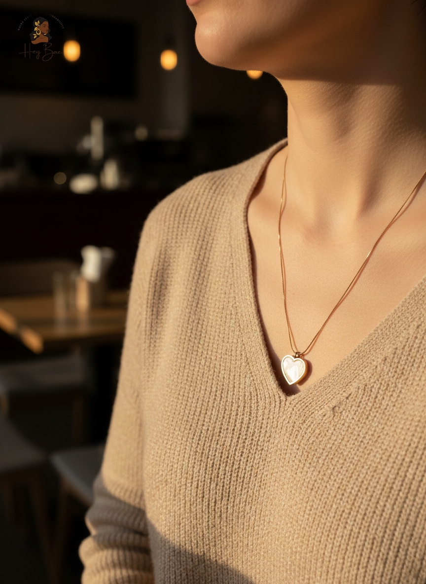 Person wearing a gold necklace with a heart pendant in a blurred indoor setting