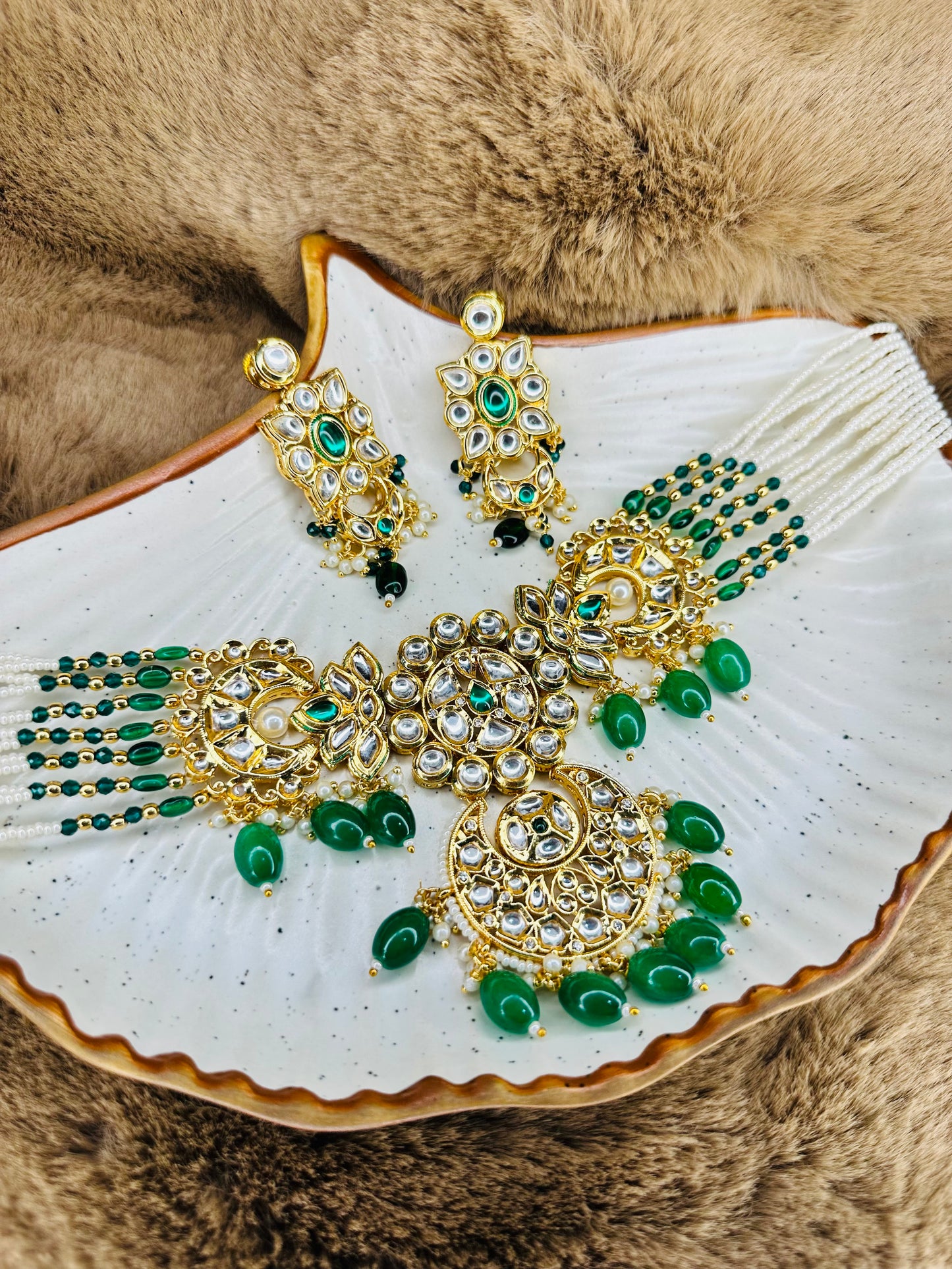A set of ethnic green kundan and pearl jewelry including a necklace and earrings, displayed on a white surface with a brown furry object in the background.