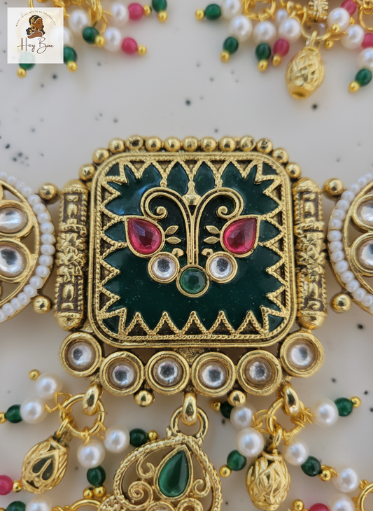 Decorative gold jewelry piece with green and red stones on a light background